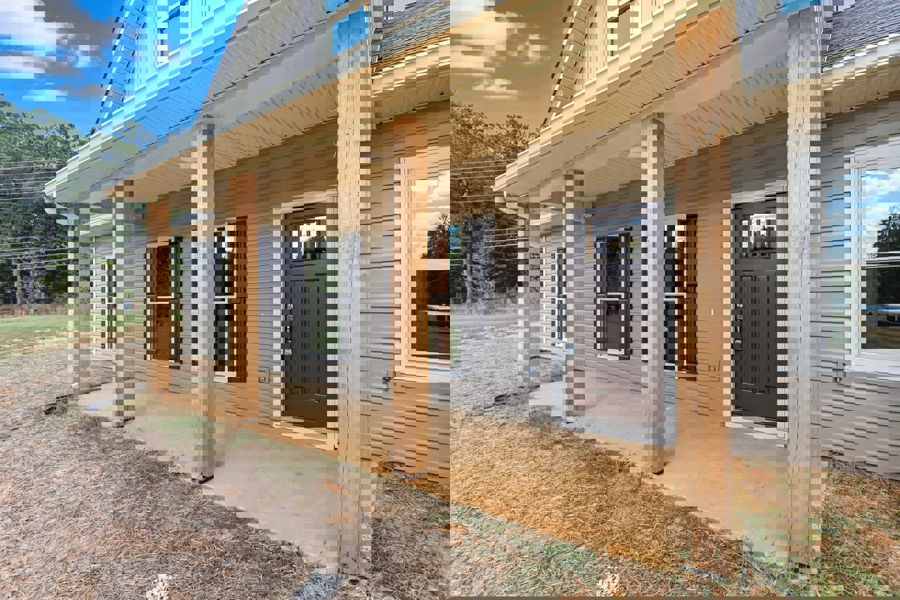 White siding house with covered porch, brown concrete steps, white framed windows, wooden front door, tree reflected in glass, patch of bare dirt beside entry.