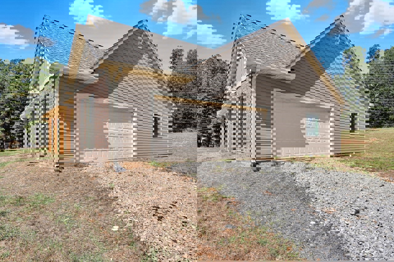 White garage door with side shadow, brick exterior with white-framed window and shutters, pile of rocks on ground, tree and plants in front yard, cloudy sky overhead