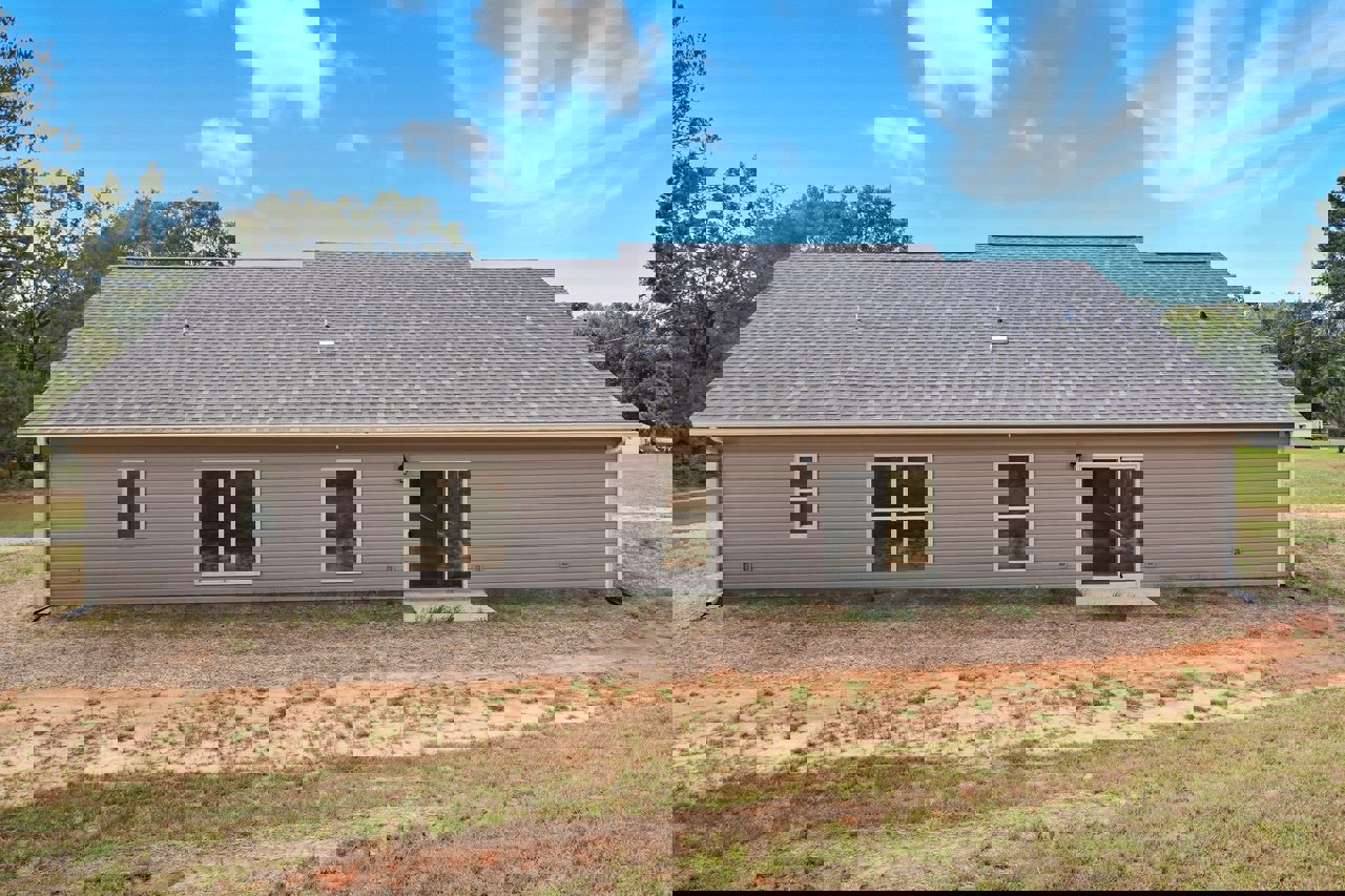 Two-story house with white-framed windows and sliding glass door, surrounded by a dirt path and patches of grass, set against a cloudy sky.