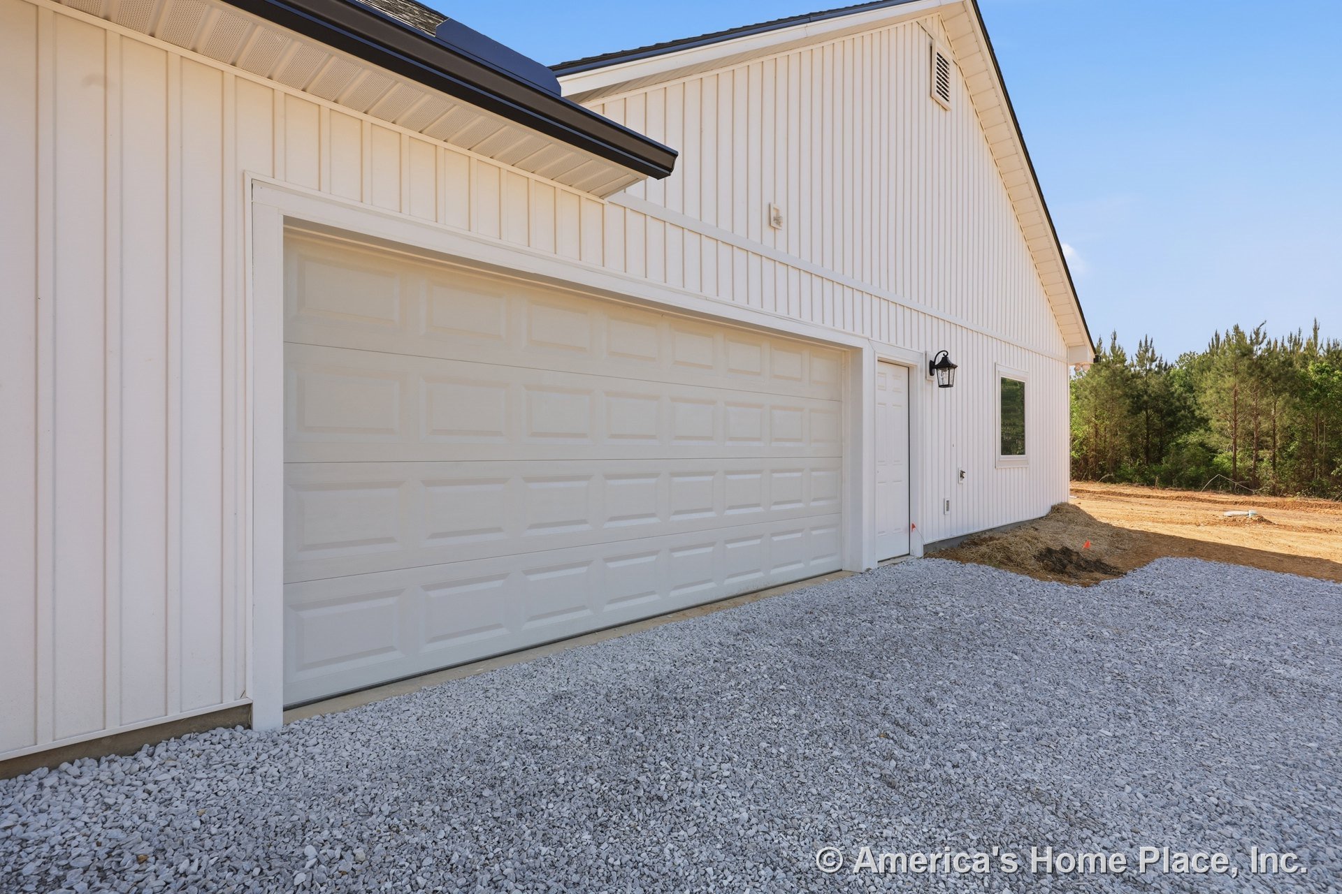 White vertical siding exterior with double paneled garage door, gravel driveway, side entry door, black outdoor wall lantern, rectangular window, white trim, and roof eaves.