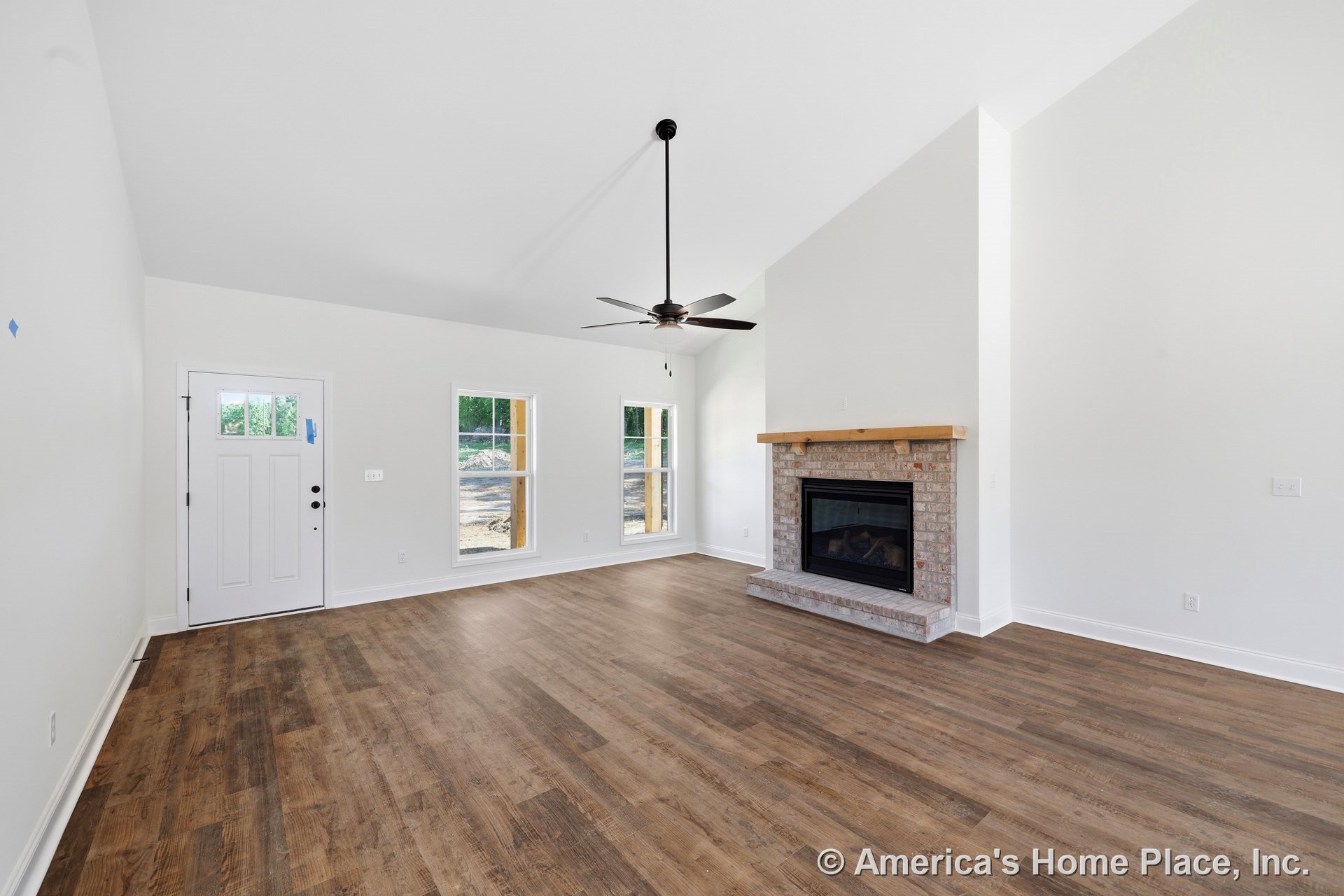 Brick fireplace with wood mantel, vaulted ceiling with ceiling fan, wide plank wood flooring, white trim and baseboards, double windows with exterior view, white front door