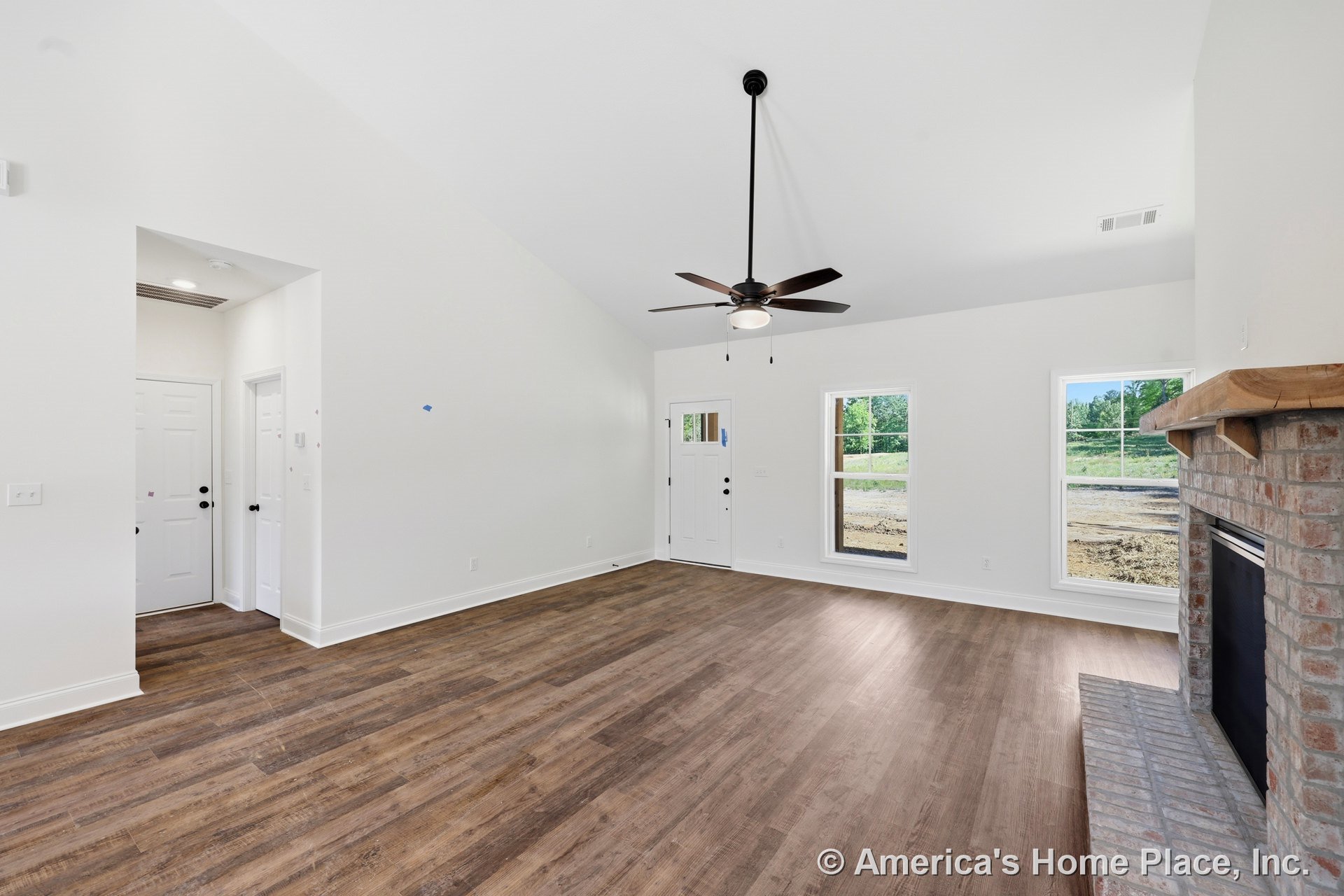 Brick fireplace with wood mantel, vaulted ceiling with ceiling fan and light, wide plank wood flooring, large windows with outdoor view, white trim and baseboards, entry door, and