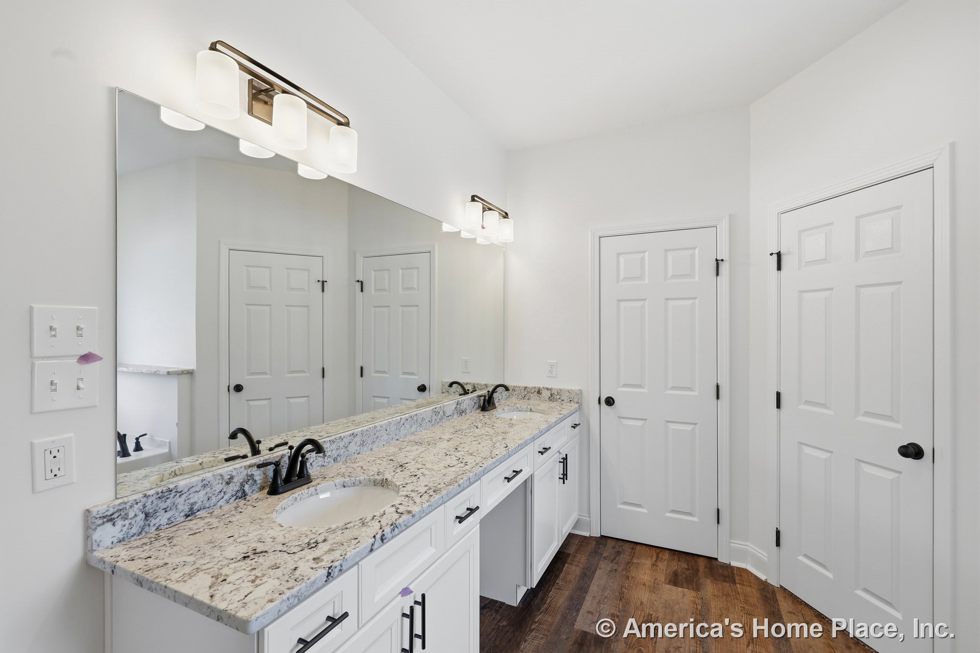 Double sink vanity with granite countertop, white shaker cabinets, large wall mirror, modern vanity lighting, black hardware, wood-look flooring, panel doors, and detailed trim.
