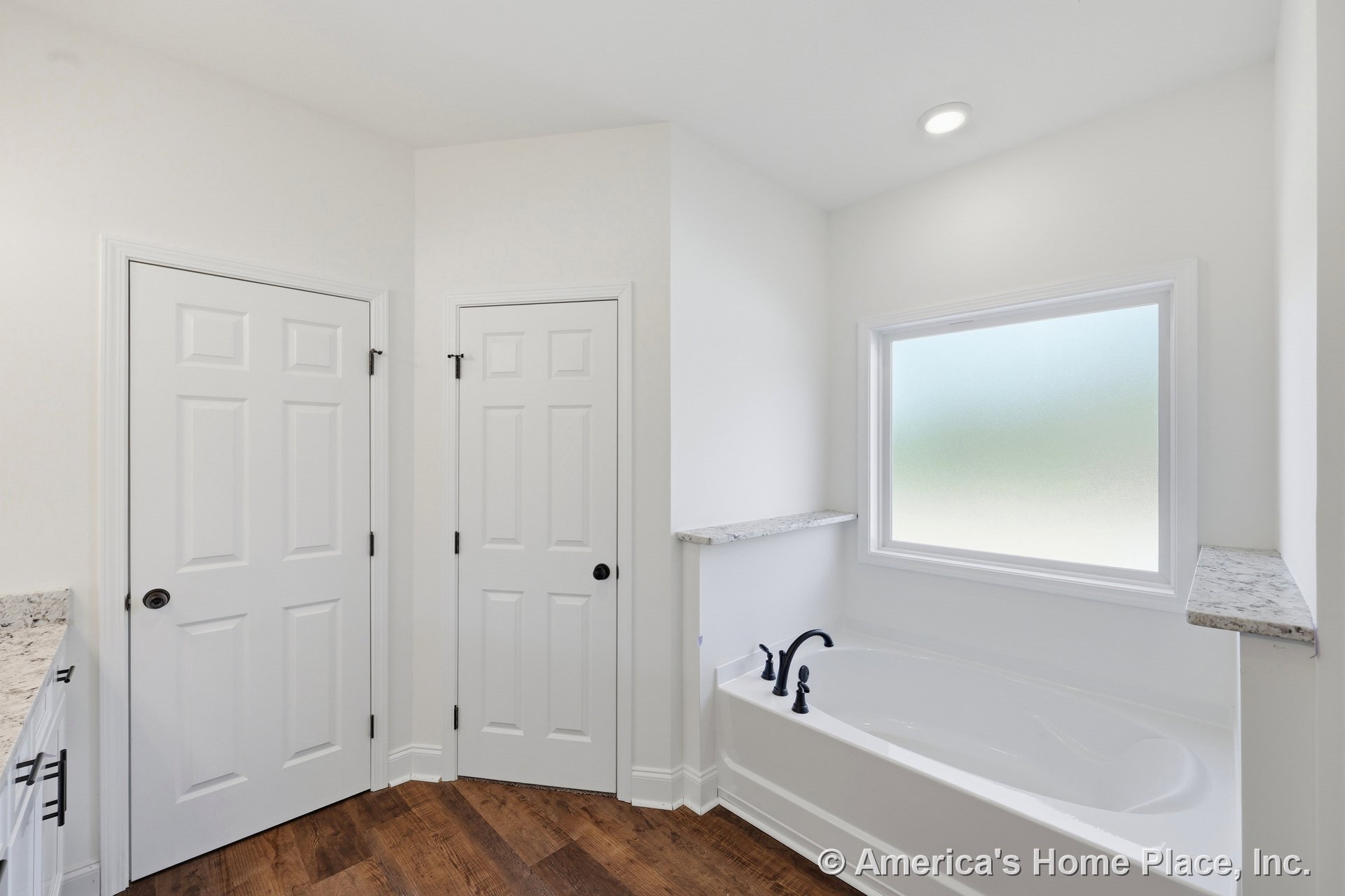 Built-in soaking tub with marble ledges and matte black faucet hardware beneath a frosted window, wood-look flooring, recessed ceiling light, white paneled doors, and built-in