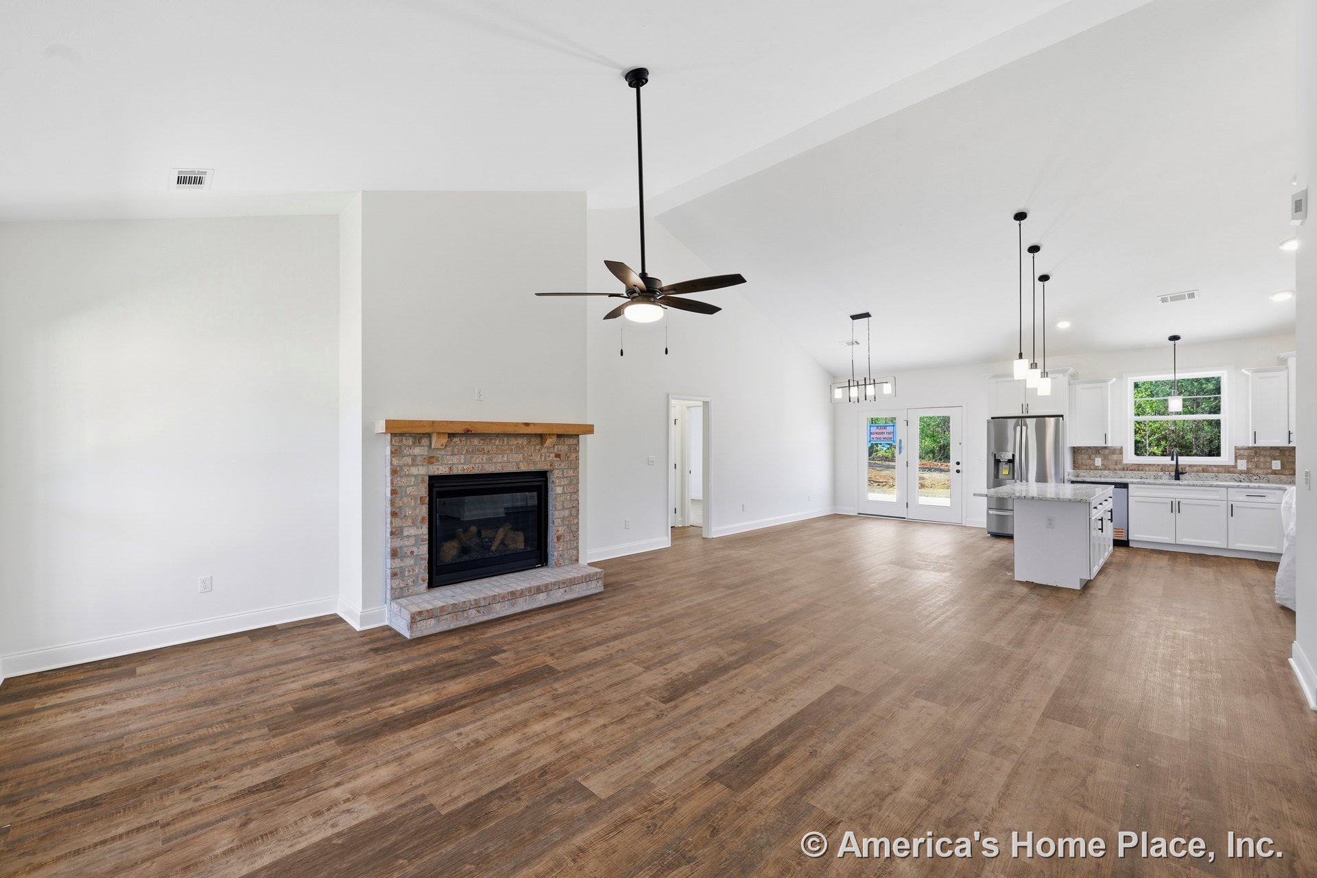 Brick fireplace with wood mantel, vaulted ceiling with ceiling fan, modern kitchen featuring white cabinetry and granite countertops, wood-look flooring throughout, pendant and