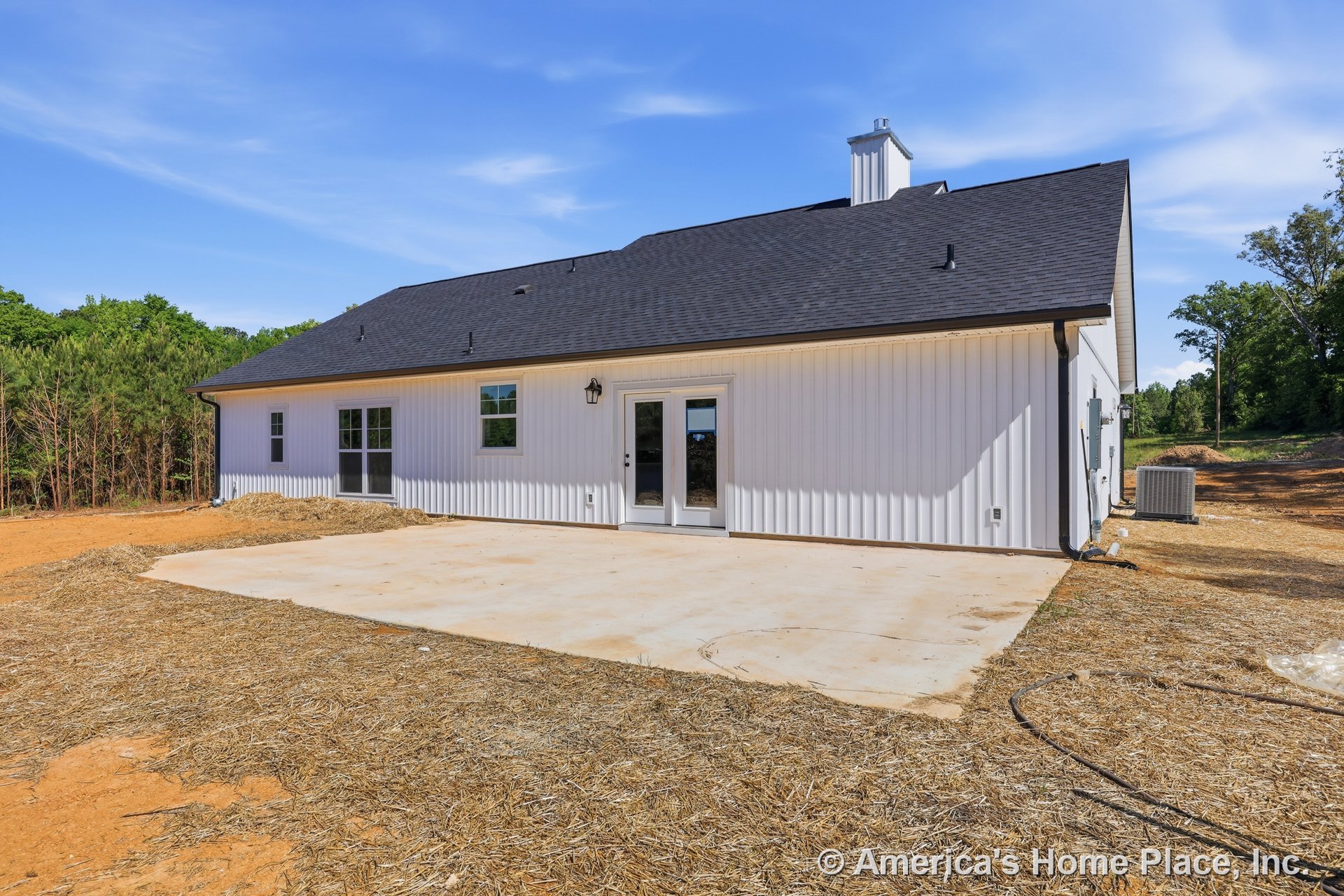 Large concrete patio adjacent to white vertical siding exterior, black shingle roof, double glass doors, multiple windows, exterior wall lantern, backyard with chimney and air