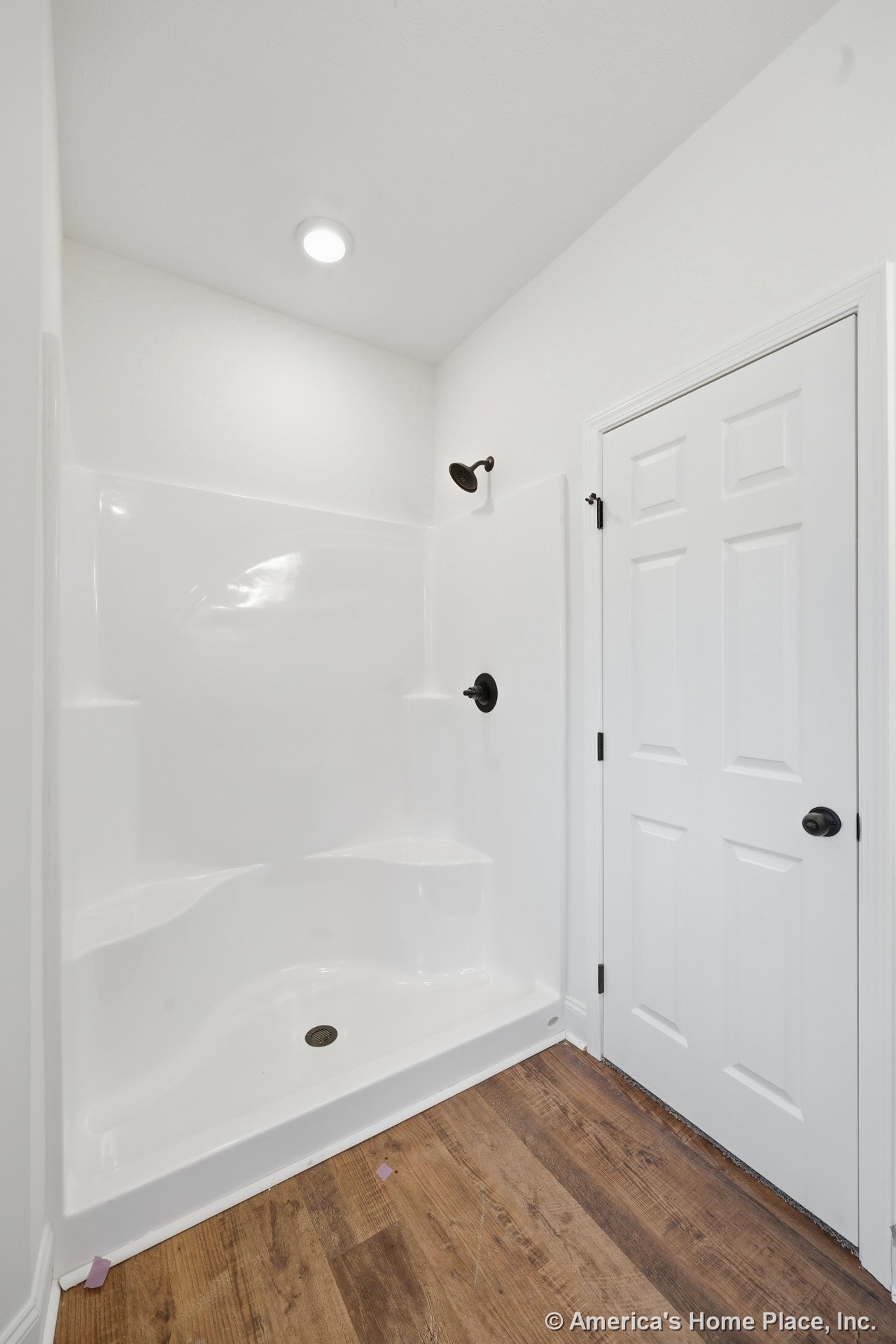 White fiberglass shower enclosure with integrated shelves and dark bronze showerhead, wood-look vinyl flooring, recessed ceiling light, and white paneled door.