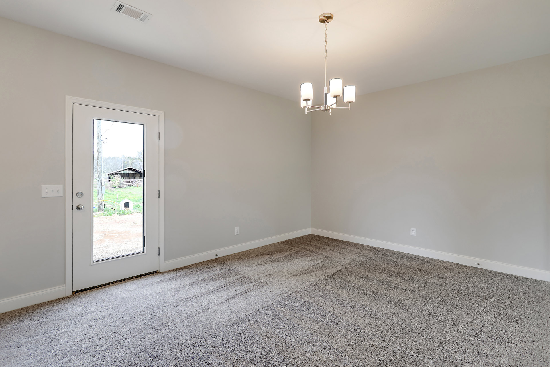 Carpeted room with white plaster walls, tile flooring near a wooden door, ornate chandelier hanging from ceiling, door opens to view of farm, shed, and barn with hay bales in
