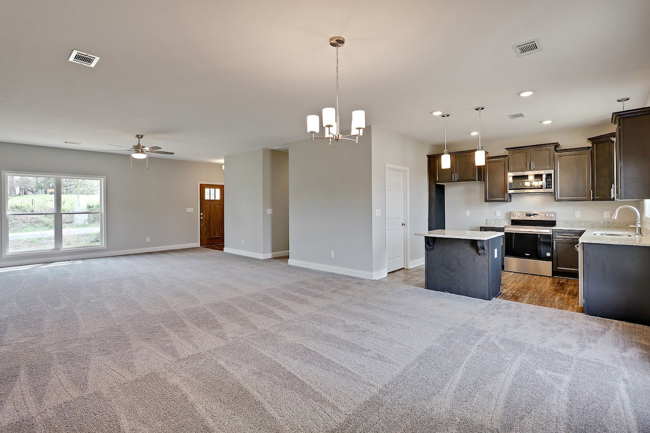 Spacious living room with carpeted floor, open kitchen featuring marble countertop and stainless steel stove, large window overlooking grassy field, white walls, and modern