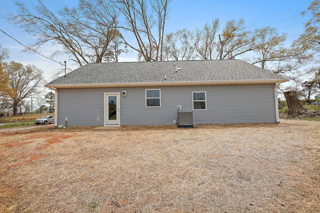 Grey siding house with white trim, front yard with green grass, mature trees in background, white front door with window, illuminated window beside entrance.