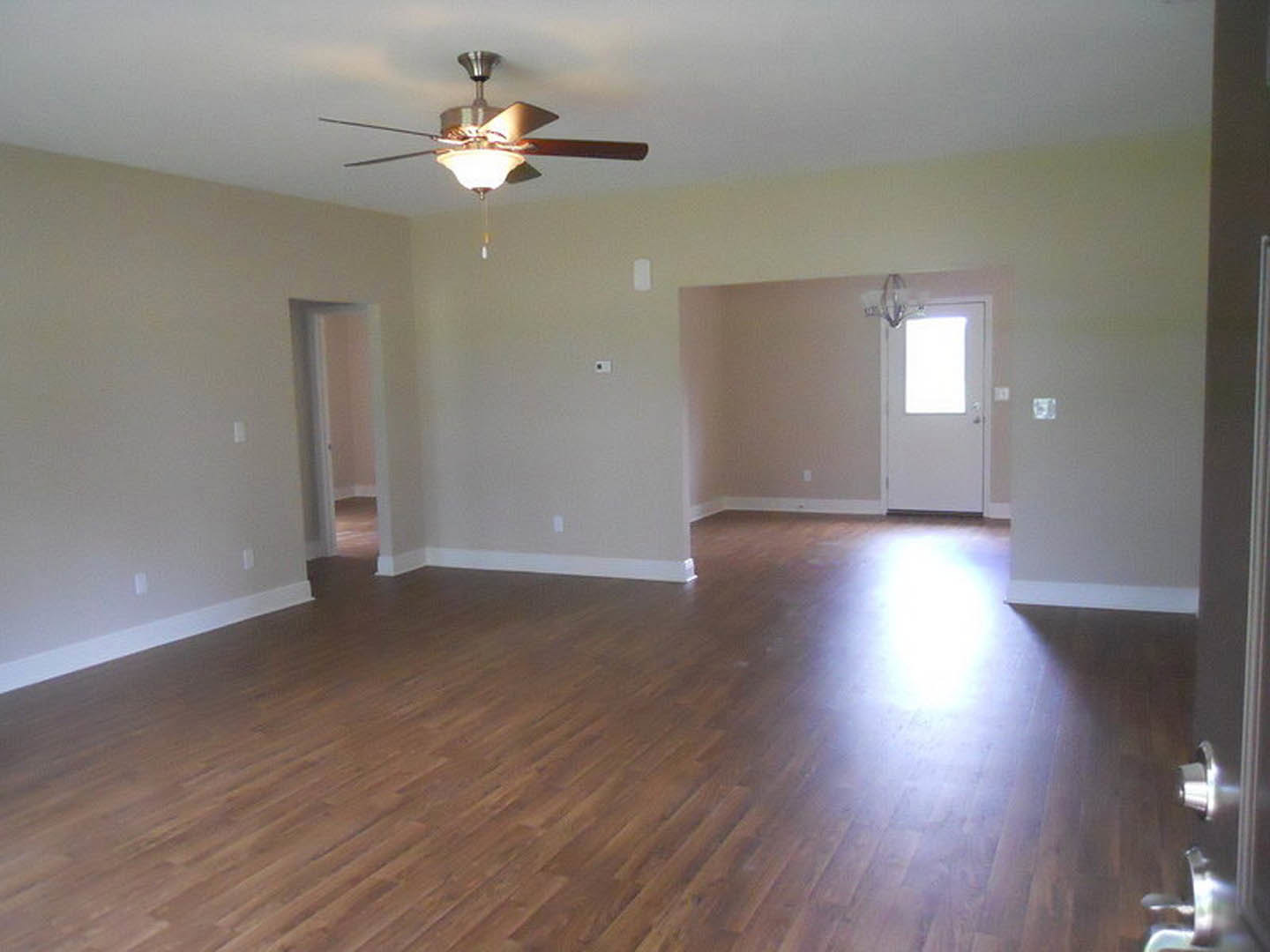 Living room with wood flooring, white walls, ceiling fan with light fixture, white door featuring glass window, rectangular black-bordered object against wall