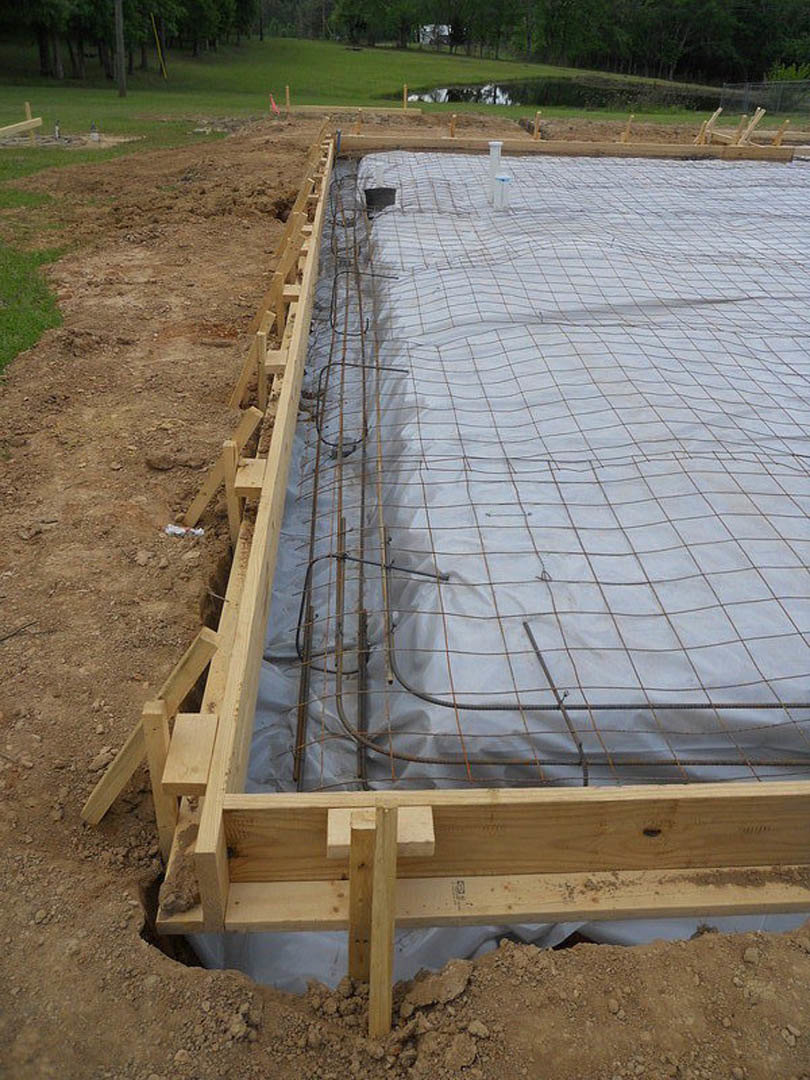 Metal rebar grid laid on concrete foundation, wooden beam and white tarp with black lines, dirt ground, trees in background