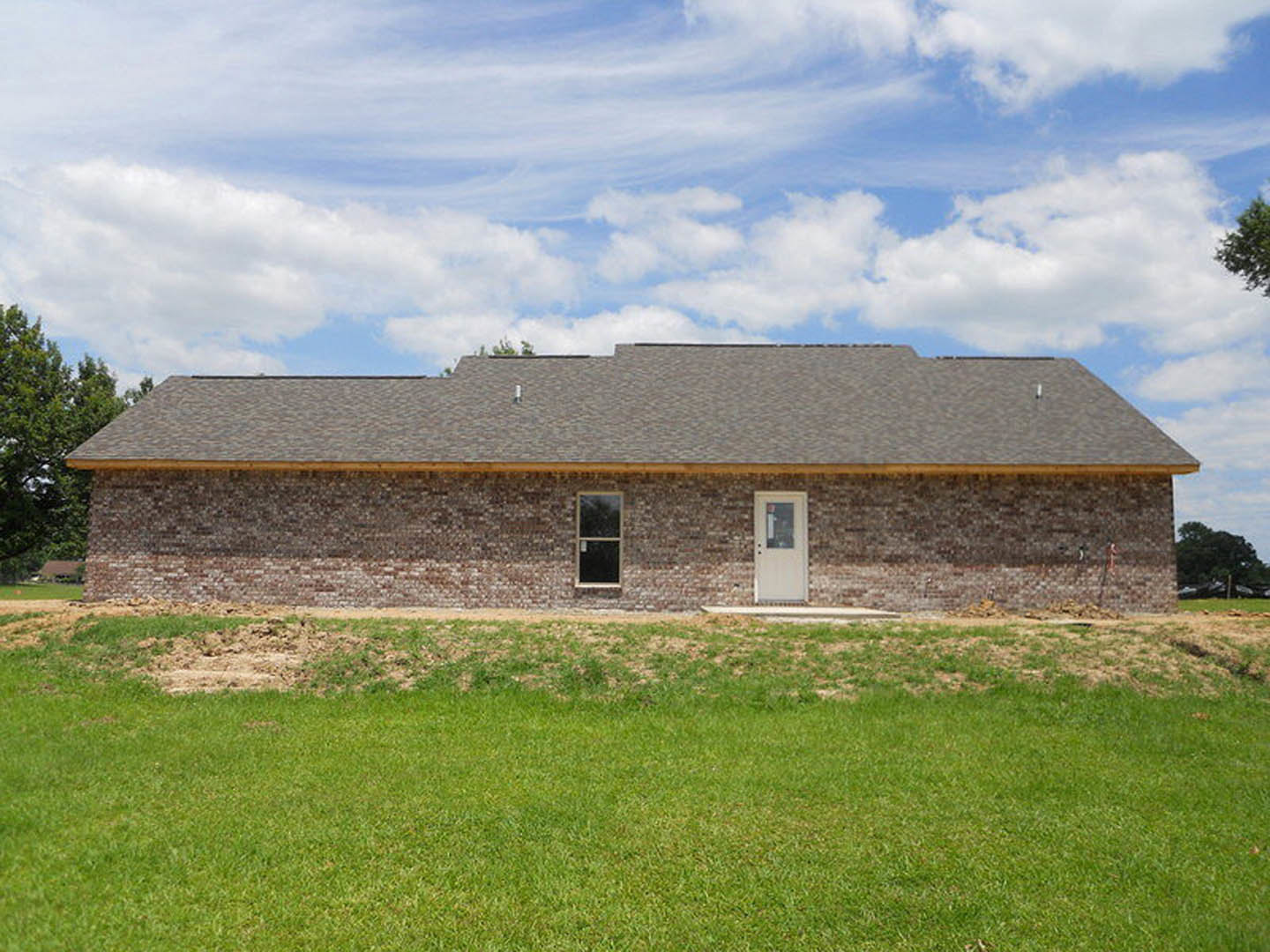 Red brick house with white front door and window, surrounded by green lawn and trees under a partly cloudy sky