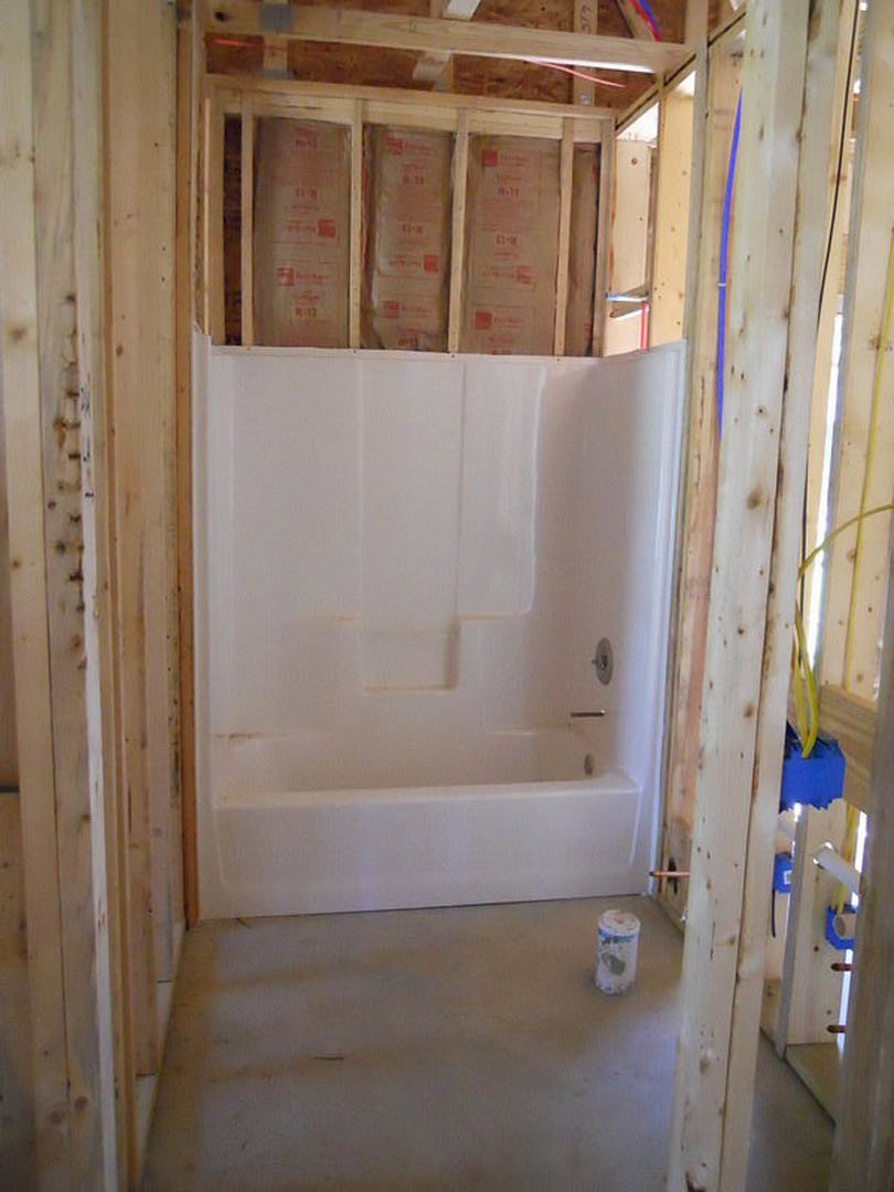 Bathroom under construction featuring a white bathtub, open door, unfinished plaster walls, exposed wood framing, and a container of paper on the floor.