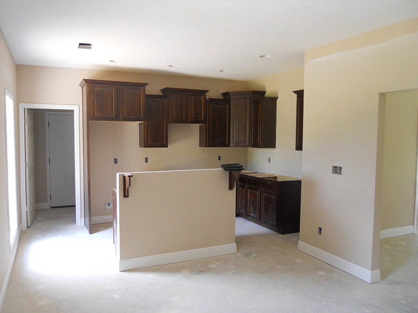 Kitchen with dark brown cabinets, white walls, wooden floating shelf, white door, and light-colored flooring with white baseboards