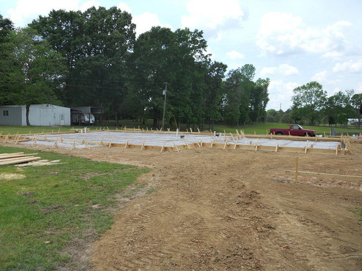Red pickup truck parked on grassy lot beside partially constructed home foundation, pile of lumber nearby, tree and fence in background under cloudy sky.