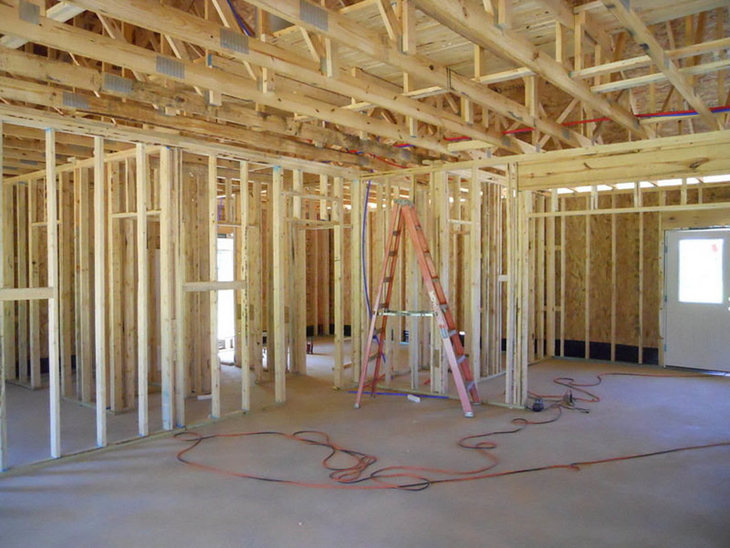 Ladder standing in unfinished room with exposed wooden ceiling beams, white door with frosted window, visible electrical wires on floor, and partially installed insulation.