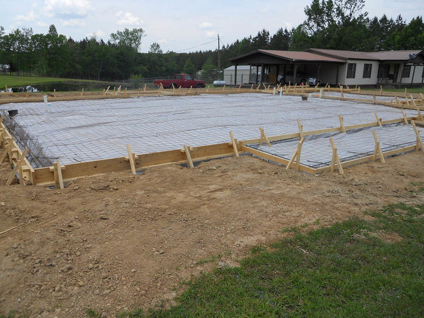 Concrete foundation and wooden framing on custom home construction site, red truck parked nearby, garage with car, dirt and grass surrounding lot, trees and clouds in background