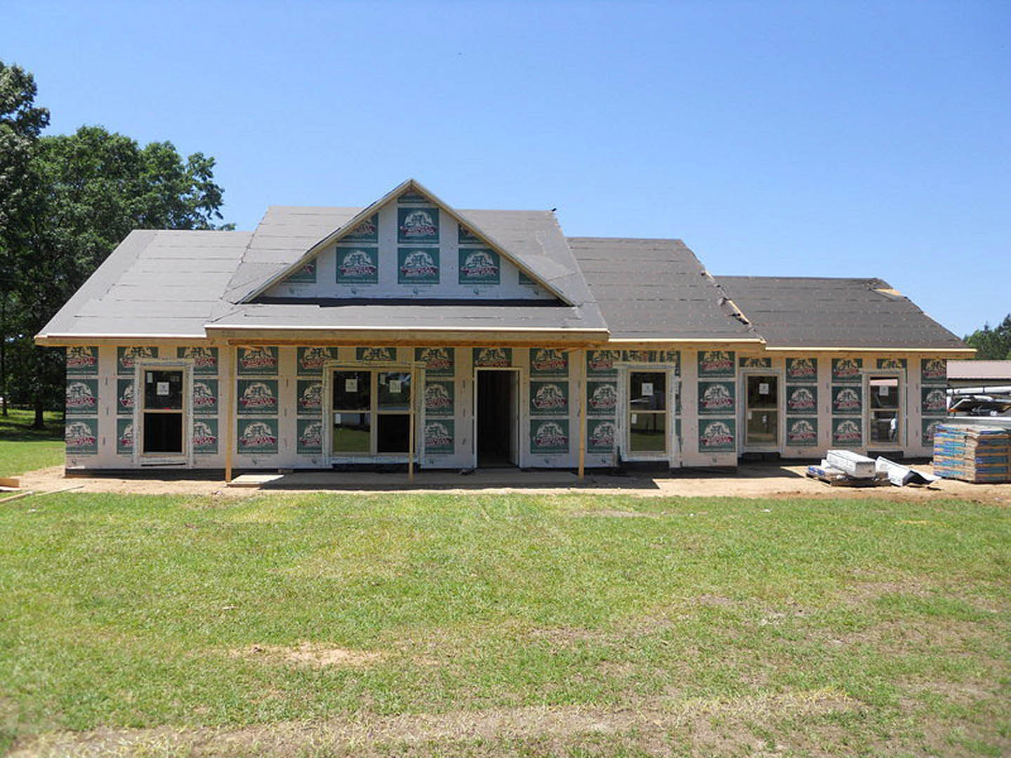 Two-story house with gray siding, white trim, covered front porch, black door, large windows, and manicured green lawn surrounded by trees under a blue sky