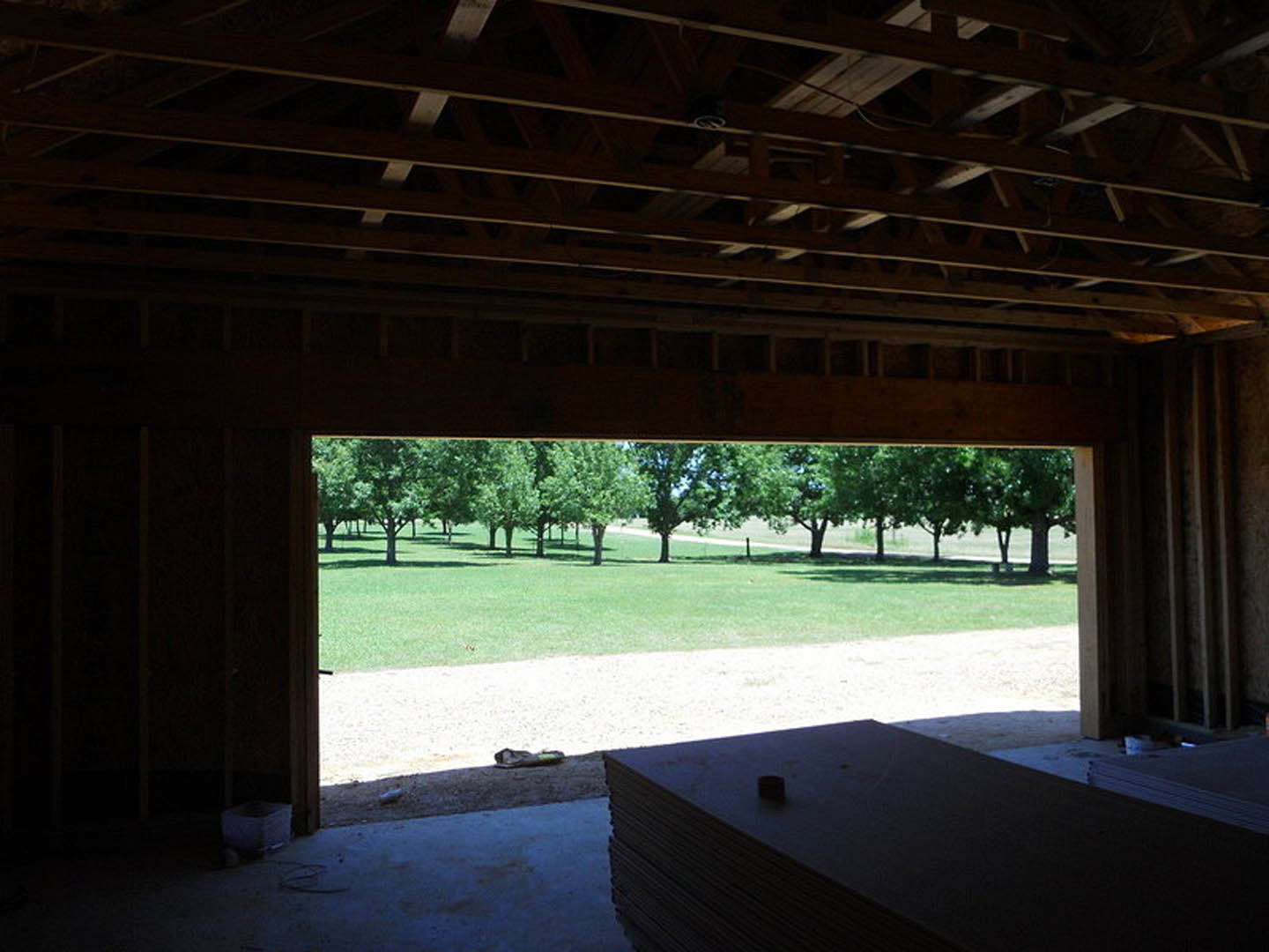 Wood-framed house under construction with exposed beams, stacks of lumber, metal materials, and surrounding grassy field with mature trees