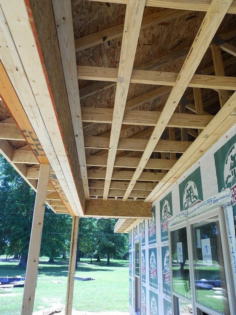 Wood-framed custom home under construction with exposed beams, unfinished wooden roof, and green lawn in foreground; leafy tree partially visible near exterior window