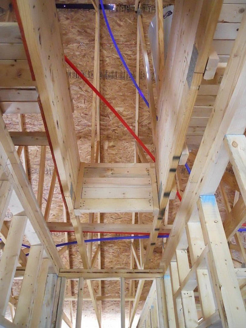 Exposed wood framing with blue and red plastic plumbing pipes, unfinished staircase, and visible insulation in a residential interior under construction