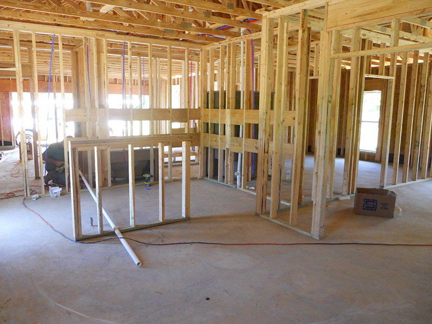 Wood-framed room under construction with exposed beams, unfinished floor, visible wiring and pipes, and a cardboard box with a blue logo on the ground.