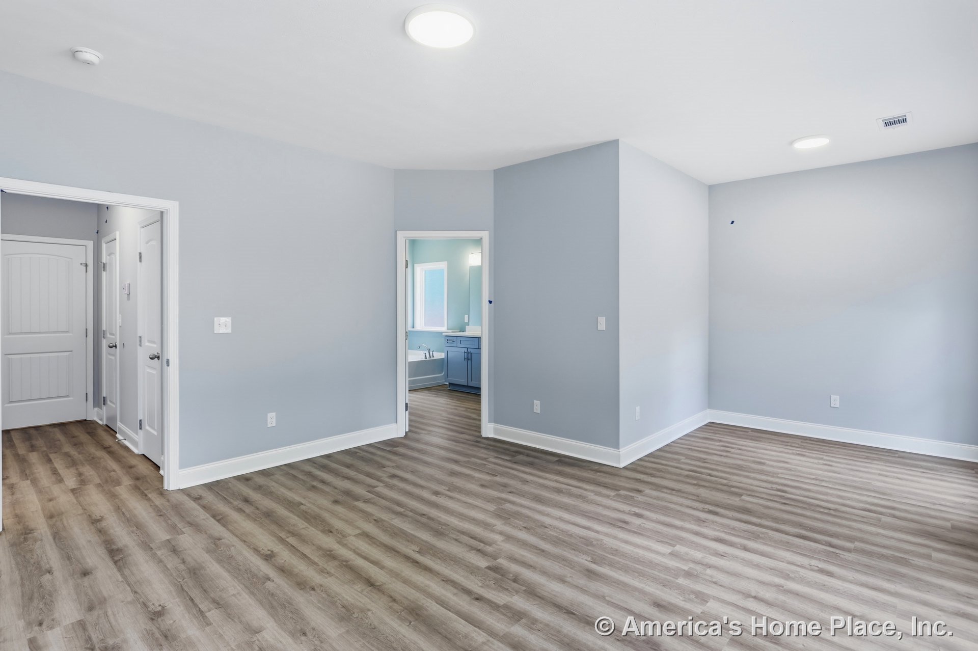 Wide open interior with light gray walls, wood-look vinyl plank flooring, white trim and baseboards, recessed ceiling lights, ceiling vent, panel doors, and bathroom cabinetry