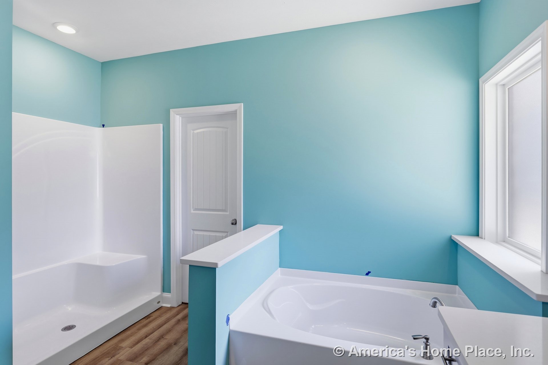 Built-in soaking tub with chrome faucet beside walk-in shower featuring molded bench, frosted window with white trim, wood-look vinyl flooring, blue painted walls, and recessed
