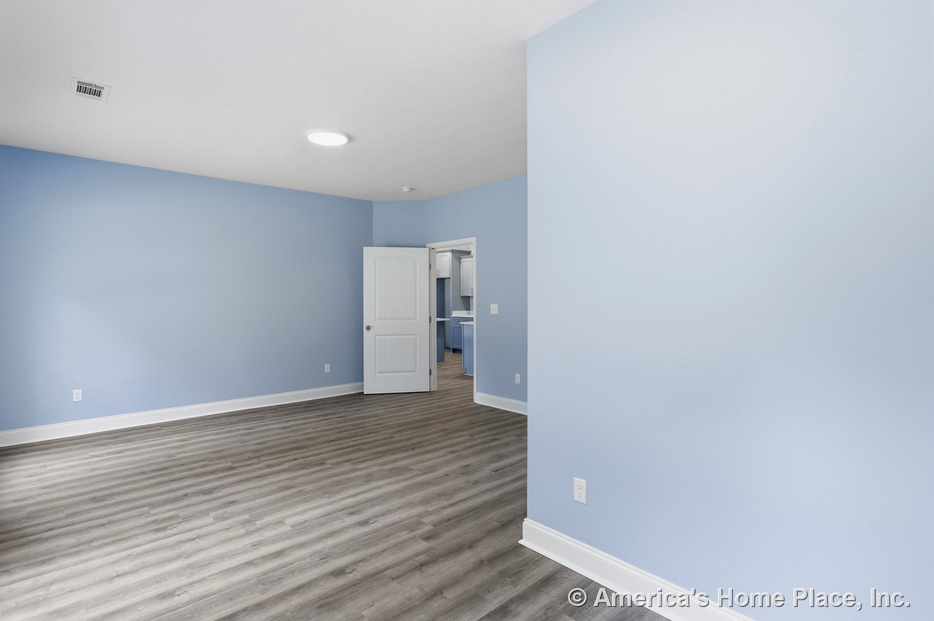 Light blue walls with white baseboards and trim, wood-look vinyl plank flooring, recessed ceiling light, paneled interior door, and open doorway revealing adjacent kitchen