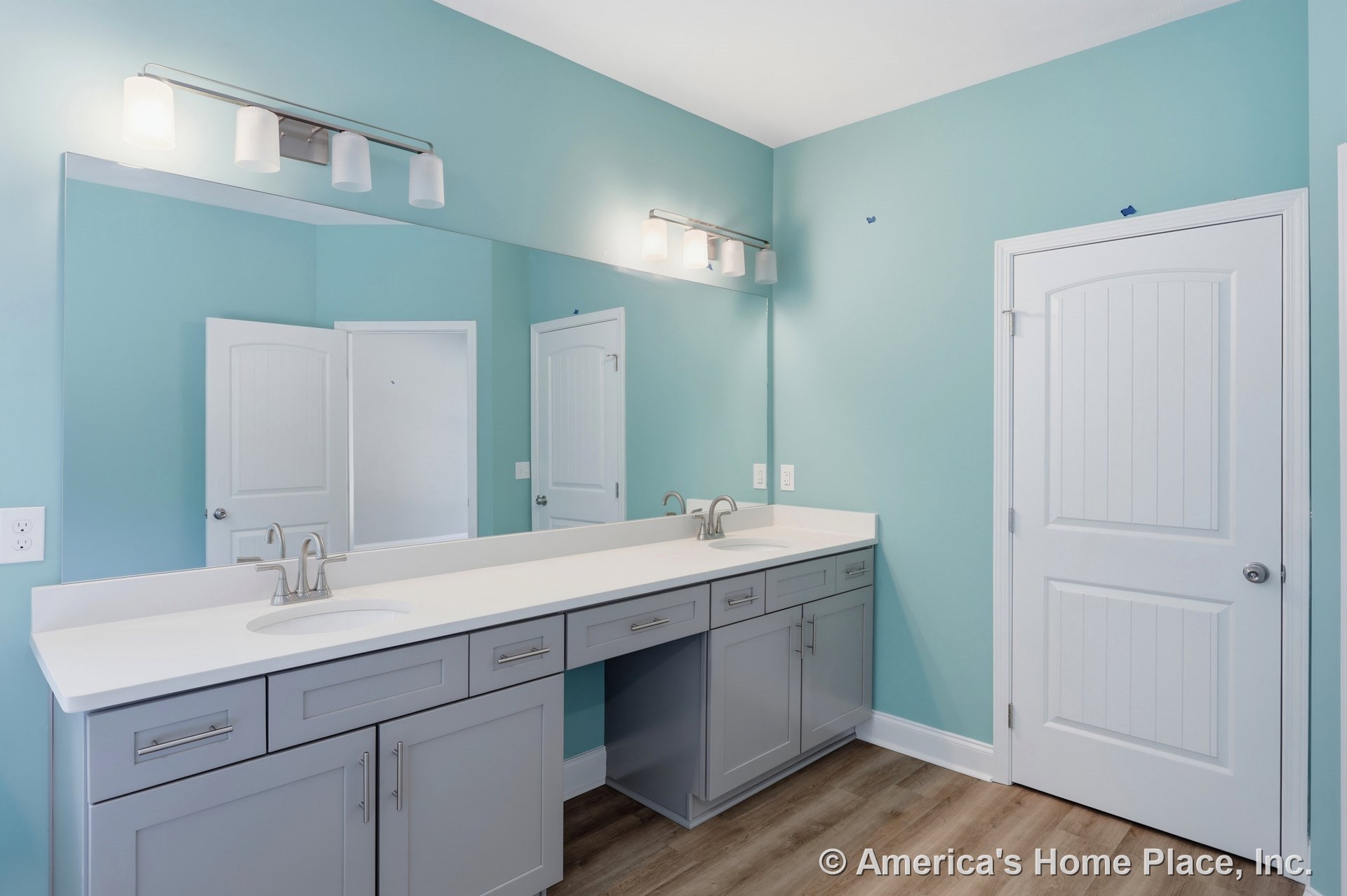 Double-sink vanity with gray shaker cabinets and white solid surface countertop, large wall mirror above, four-light vanity fixture, wood-look vinyl flooring, painted walls with