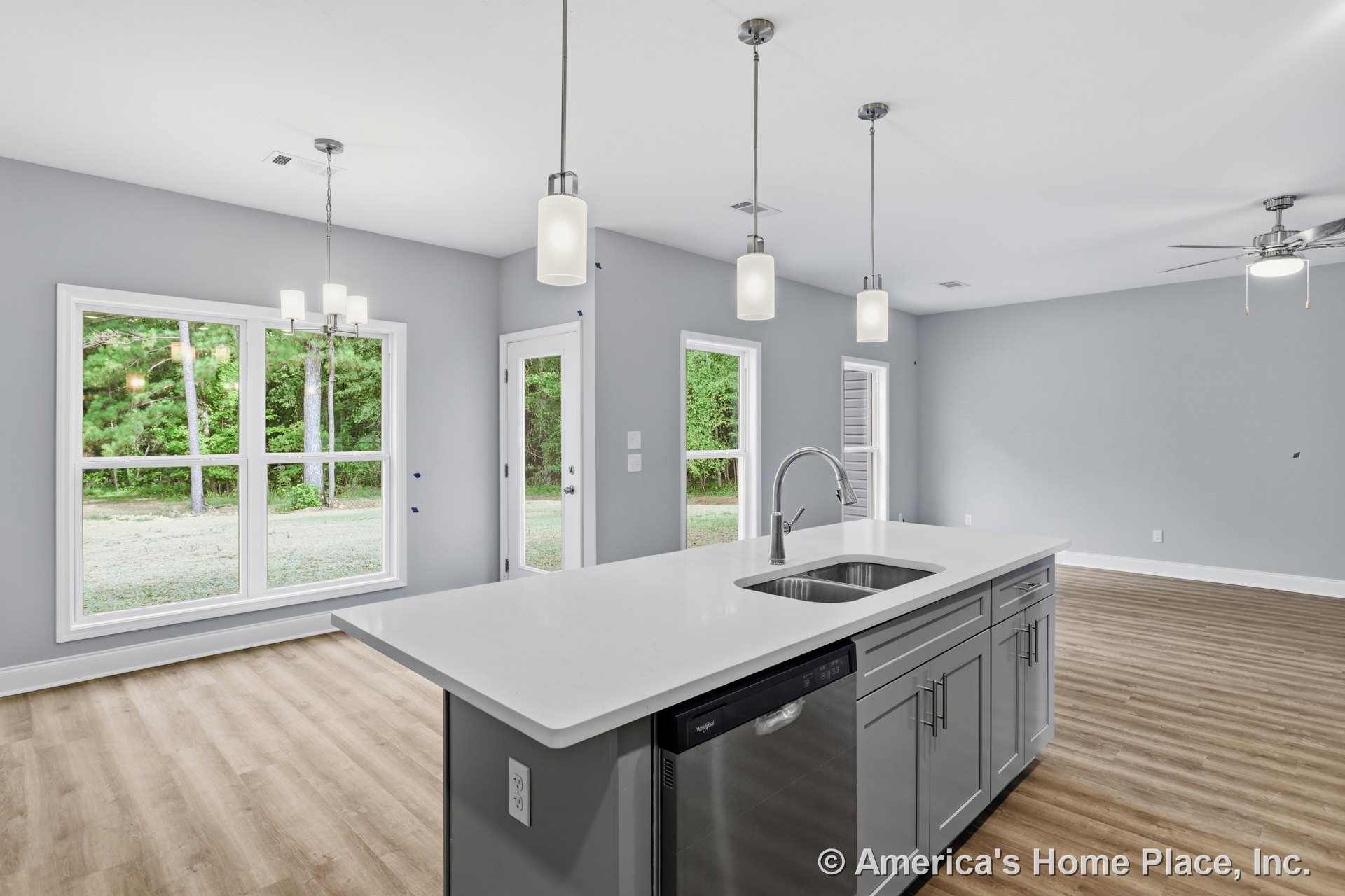 Spacious kitchen with quartz island, gray shaker cabinets, pendant lights, and large windows with white trim, opening to living area with light wood plank flooring and ceiling fan.