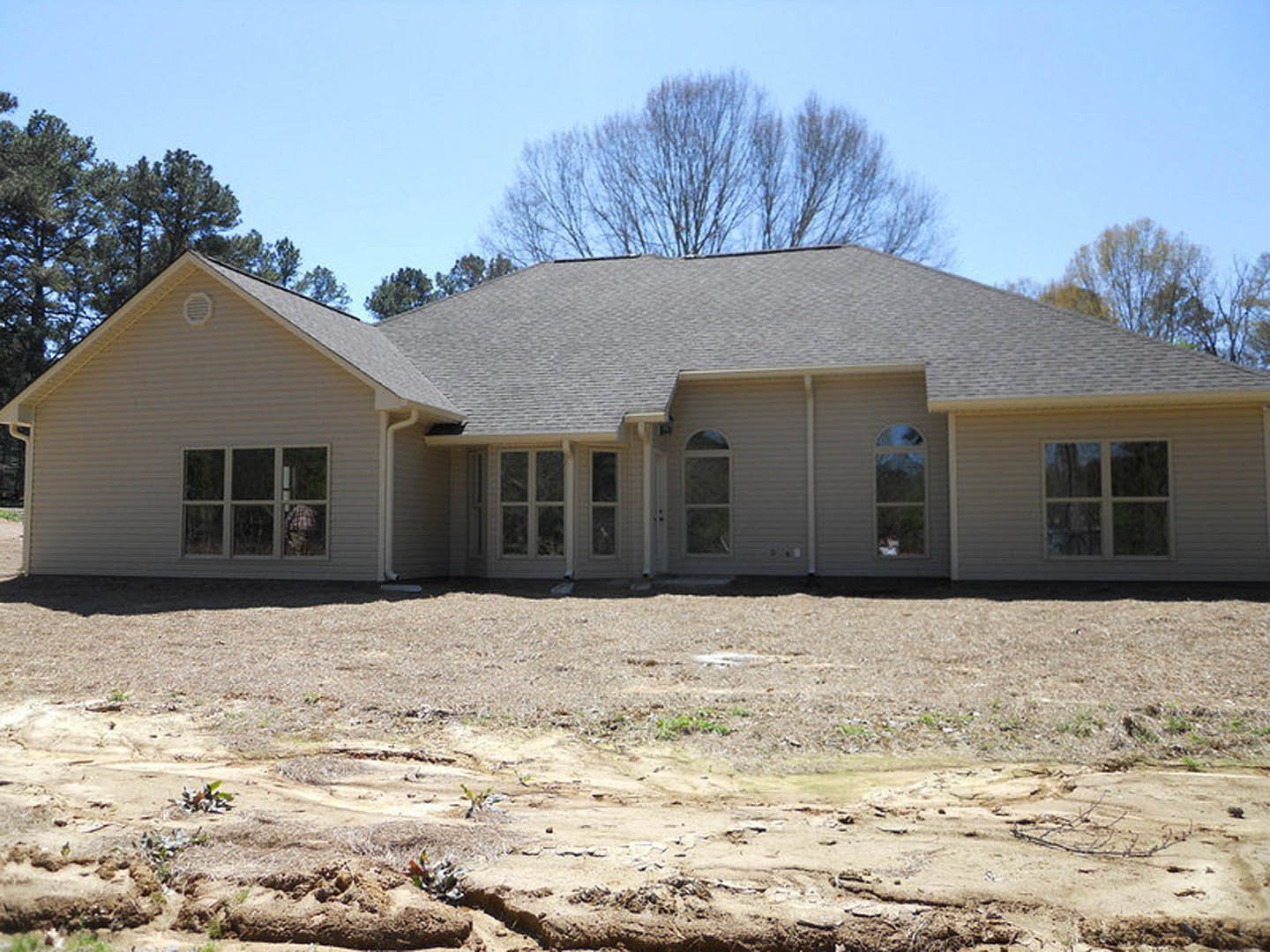 Two-story house with gray siding and white-framed windows, surrounded by bare trees and large areas of exposed dirt in the yard