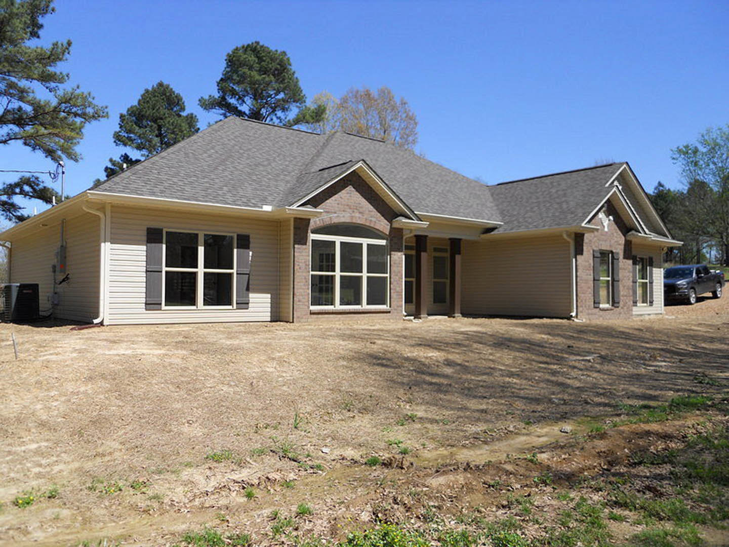 Two-story home with light siding, large white-framed windows, dirt hill and patch in front yard, black truck parked on road, trees in background