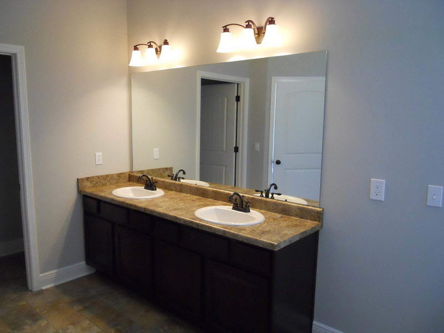 Bathroom with two white sinks set in a stone countertop, large frameless mirror above, chrome faucets, tiled backsplash, wall outlet and light switch visible, soft overhead