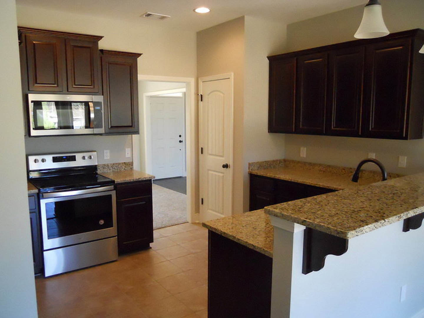 Granite countertops and dark wood cabinets in a modern kitchen with stainless steel stove, window above the sink, pendant lamp shade, and paneled cabinet doors