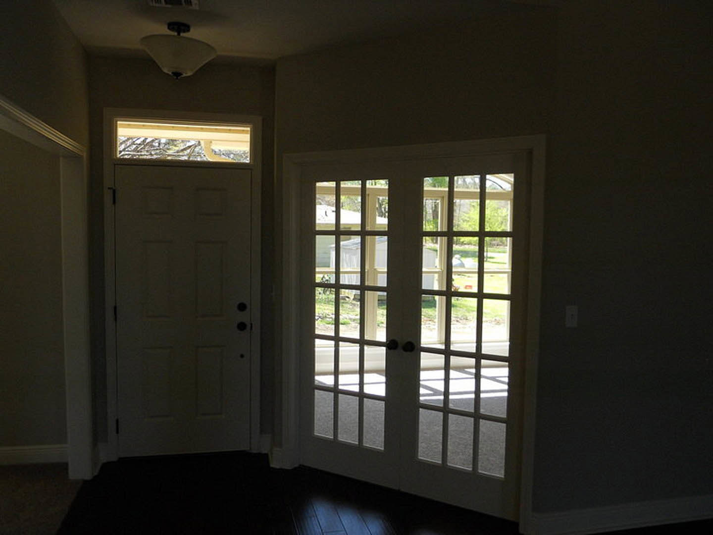 White double doors with glass panes set against a dark wall, light fixture with white shade overhead, hardwood flooring, window allowing daylight into the room.