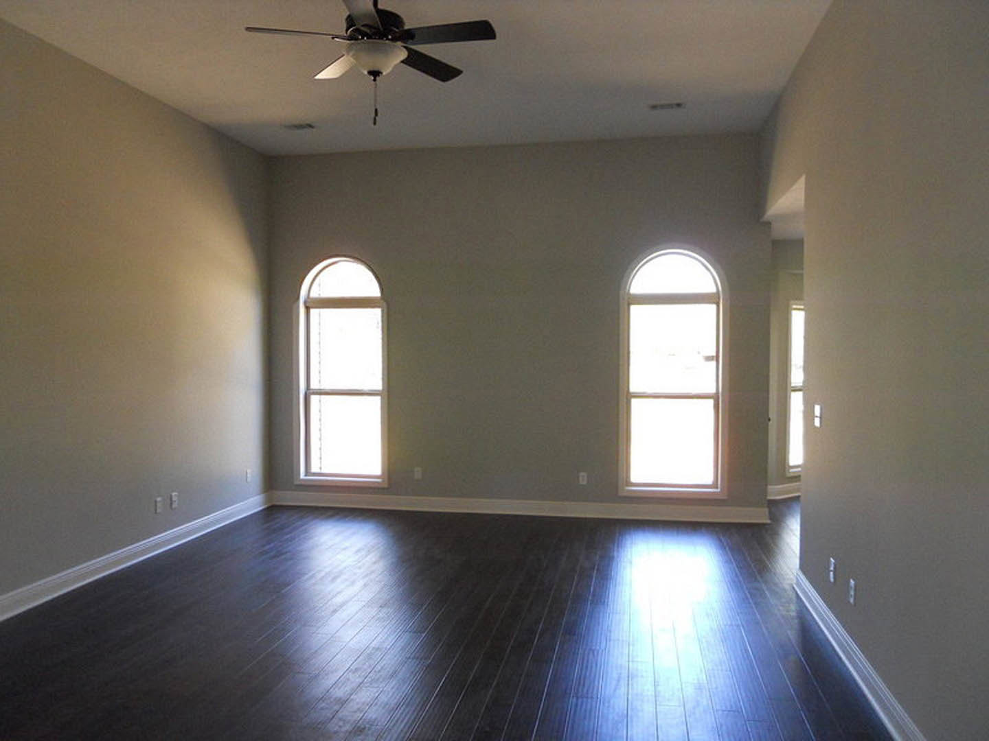 Spacious room with two arched windows, dark wood flooring, ceiling fan with light fixture, white walls, natural daylight illuminating the floor