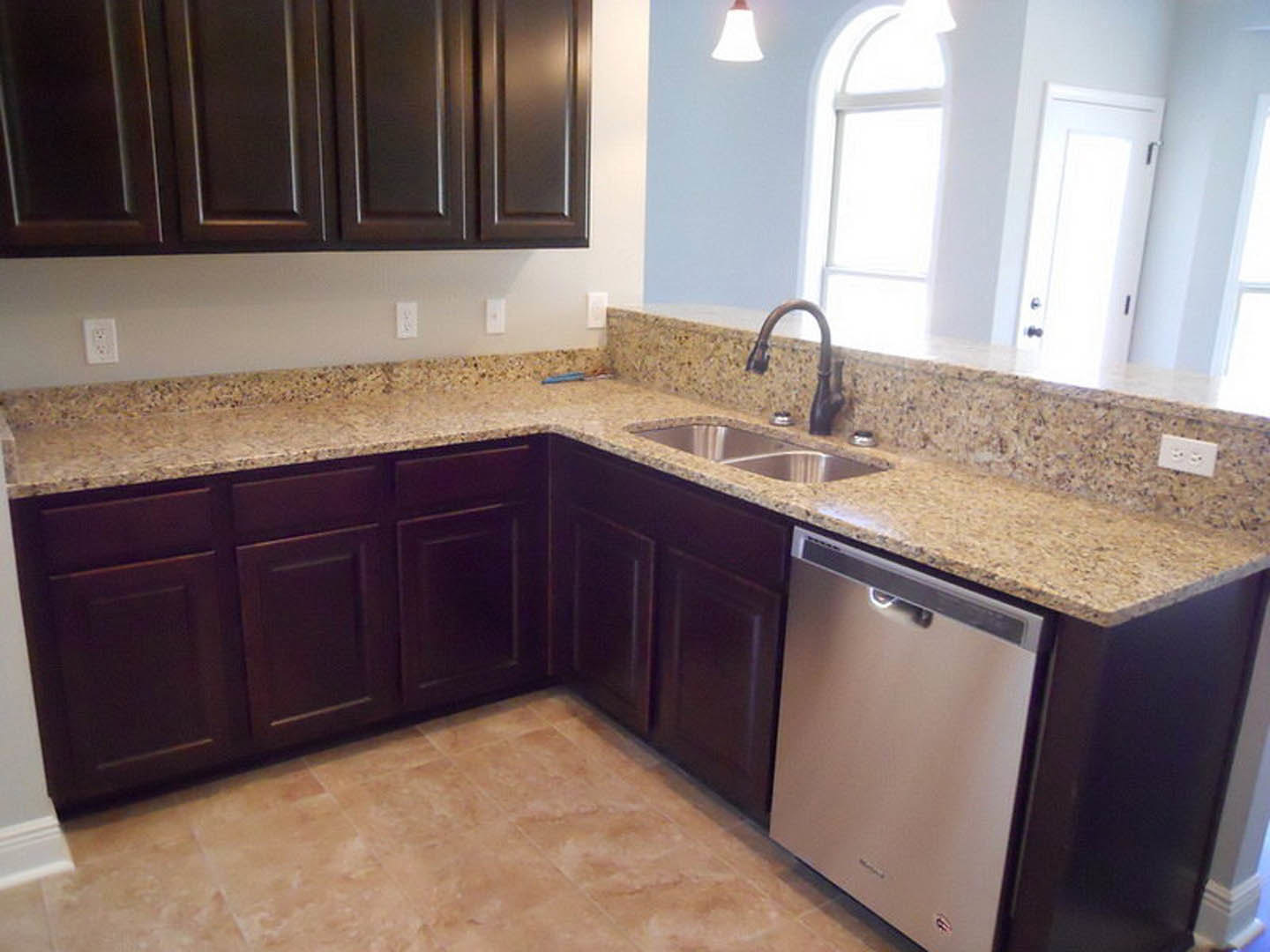 Granite countertops with dark brown cabinets, stainless steel faucet, tile backsplash, and built-in dishwasher in a modern kitchen