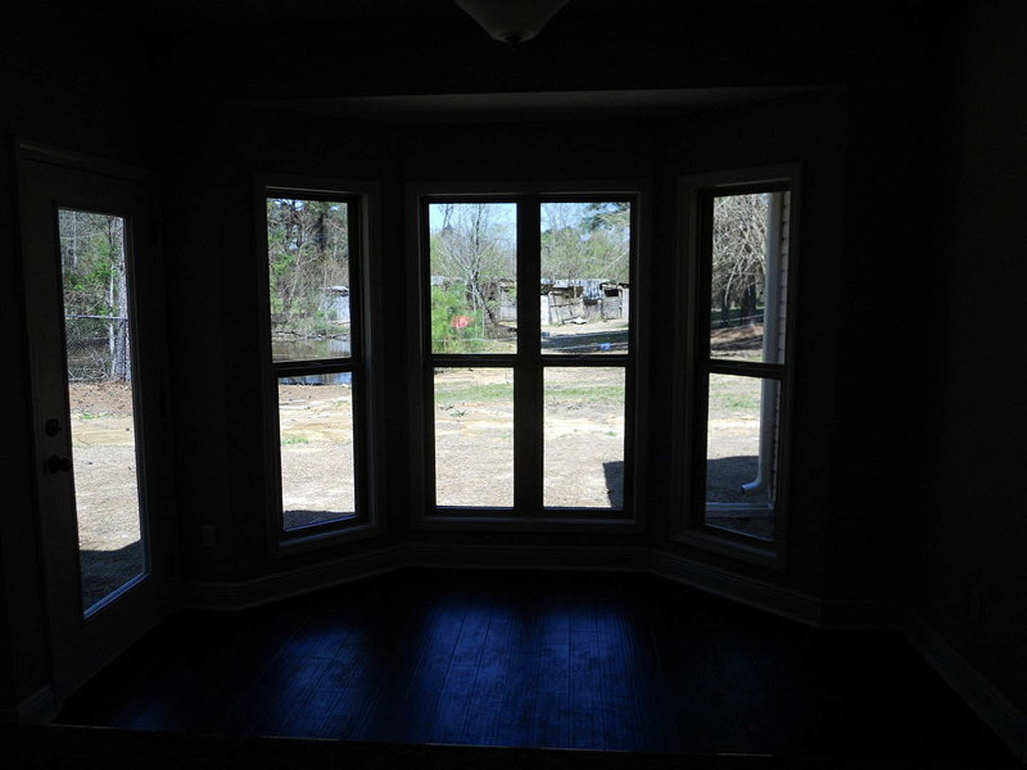 Large window framed by white trim, overlooking green grass, a fence, and a distant building; sunlight brightens the room’s neutral walls and blue accent surface.