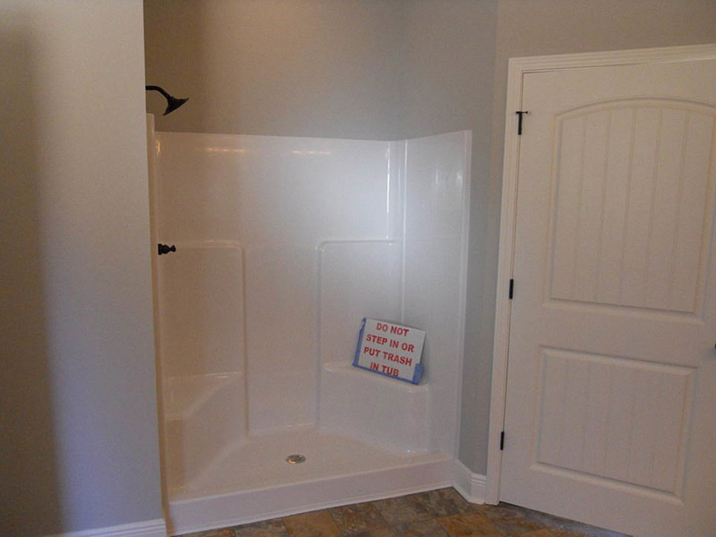 White walk-in shower with glass door, black handle, and wall-mounted sign; smooth plaster walls and light tile flooring in modern bathroom.