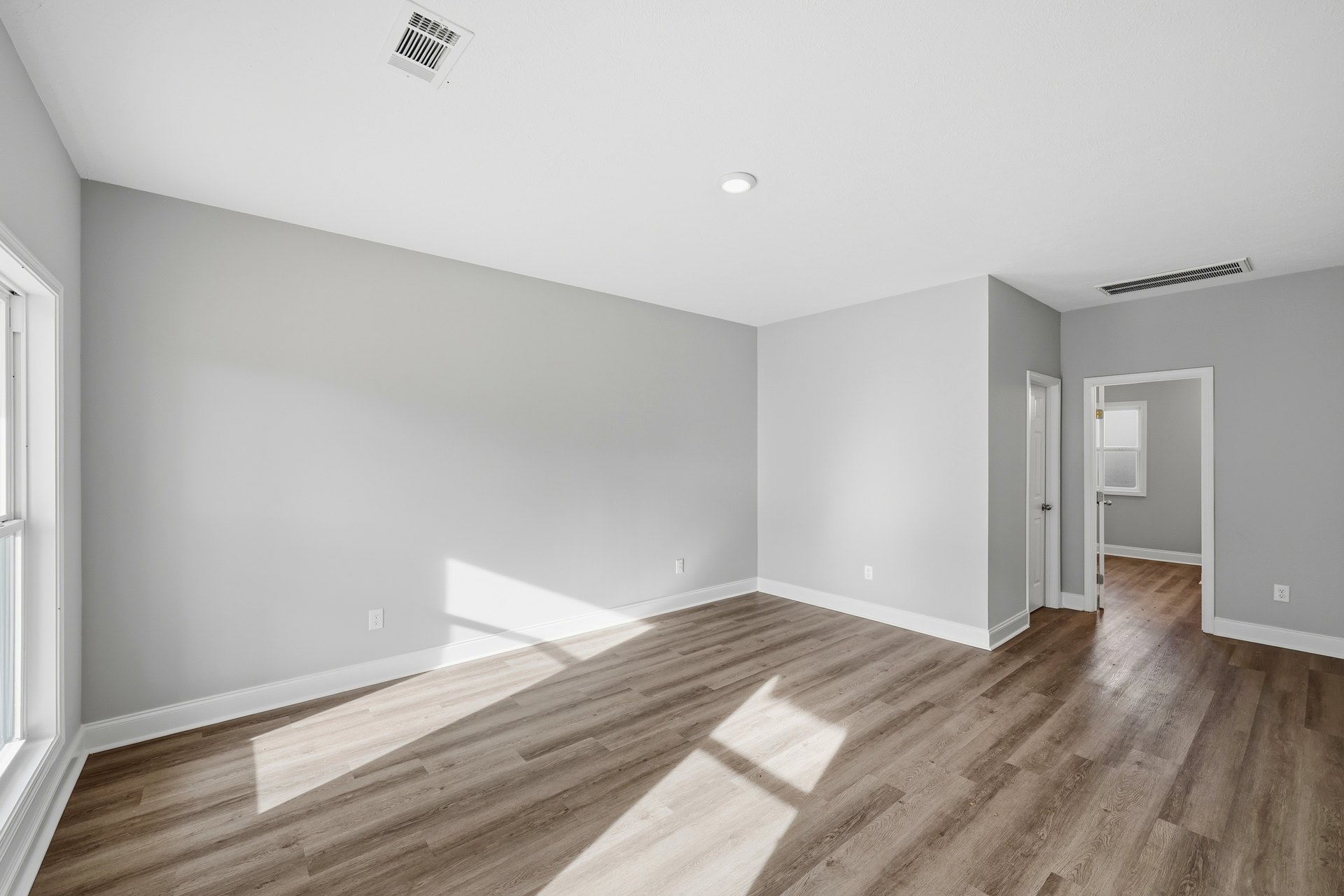 Wood flooring in a room with white plaster walls, ceiling vent, open door leading to a window, and natural light illuminating the space