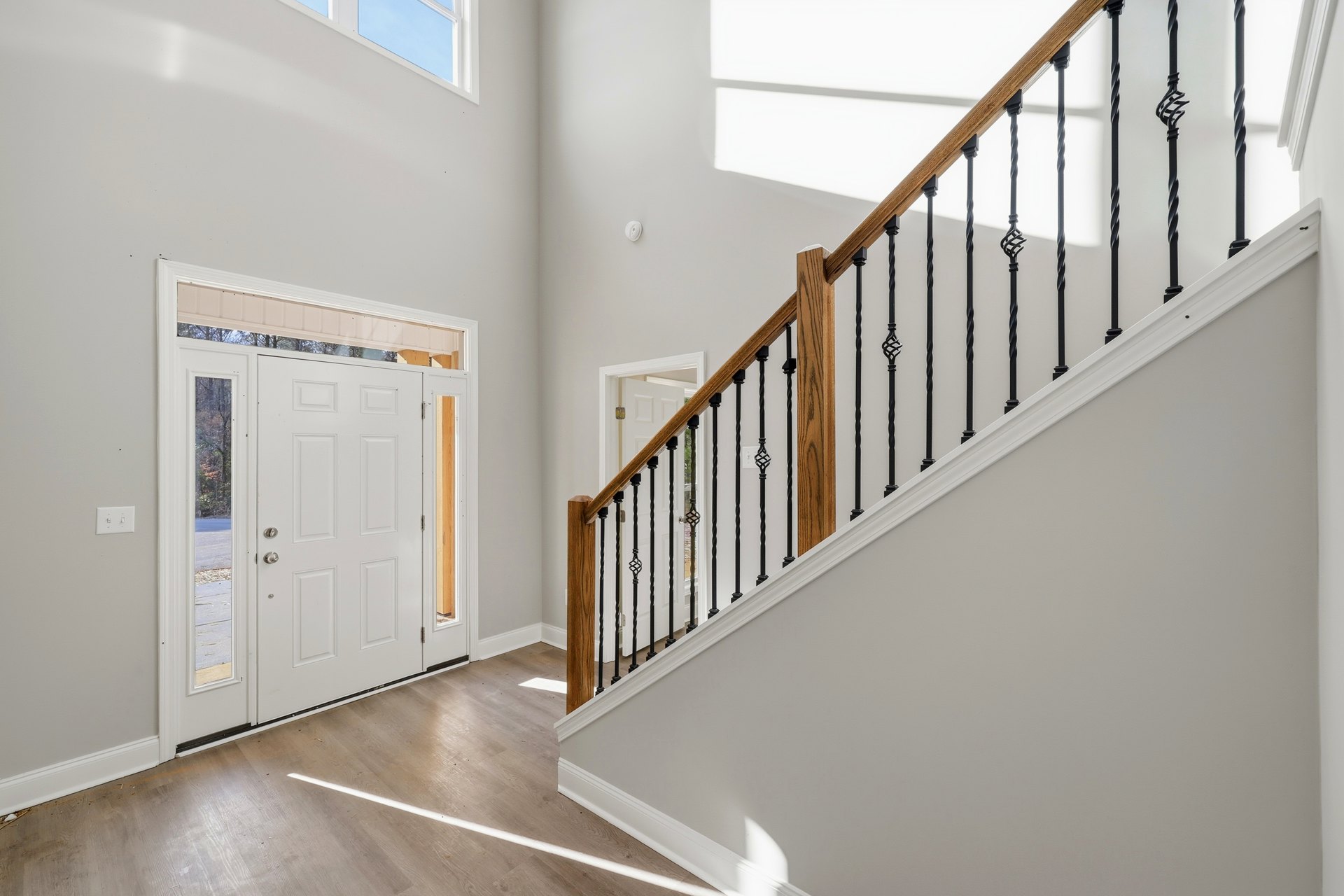 Wood staircase with black metal railing, white plaster walls, white door with glass window, and light wood flooring with white trim