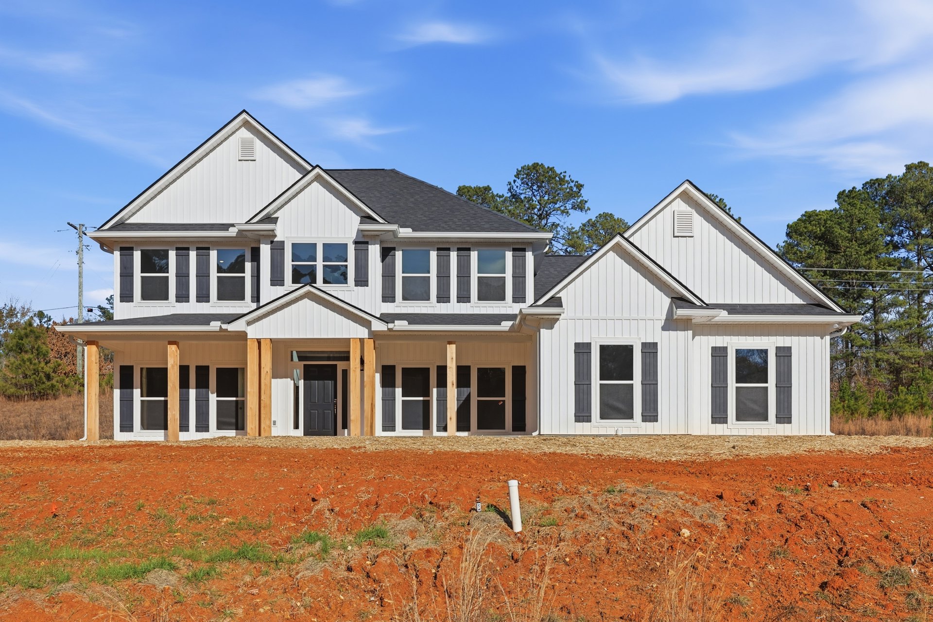 Two-story house with large front porch, white windows featuring red stickers, expansive front yard with exposed white pipe in dirt, mature trees in background, partly cloudy sky