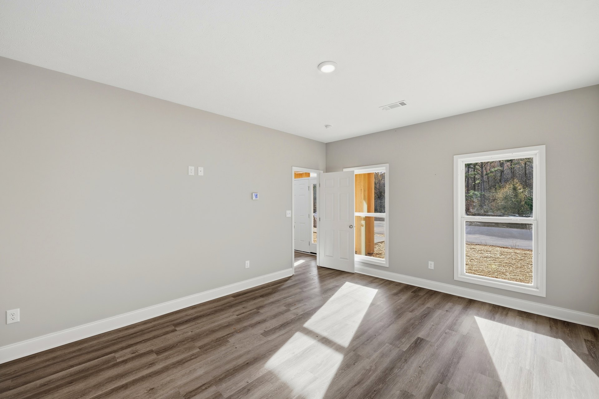 Wood flooring in a bright room with white plaster walls, a glass-paneled door, and a large window overlooking trees and grass, sunlight streaming across the floor.