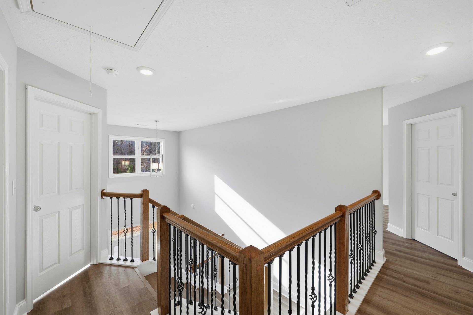 Wood staircase with black metal balusters, wooden handrail, laminate flooring, white doors with silver hardware, large window framing leafy trees outside