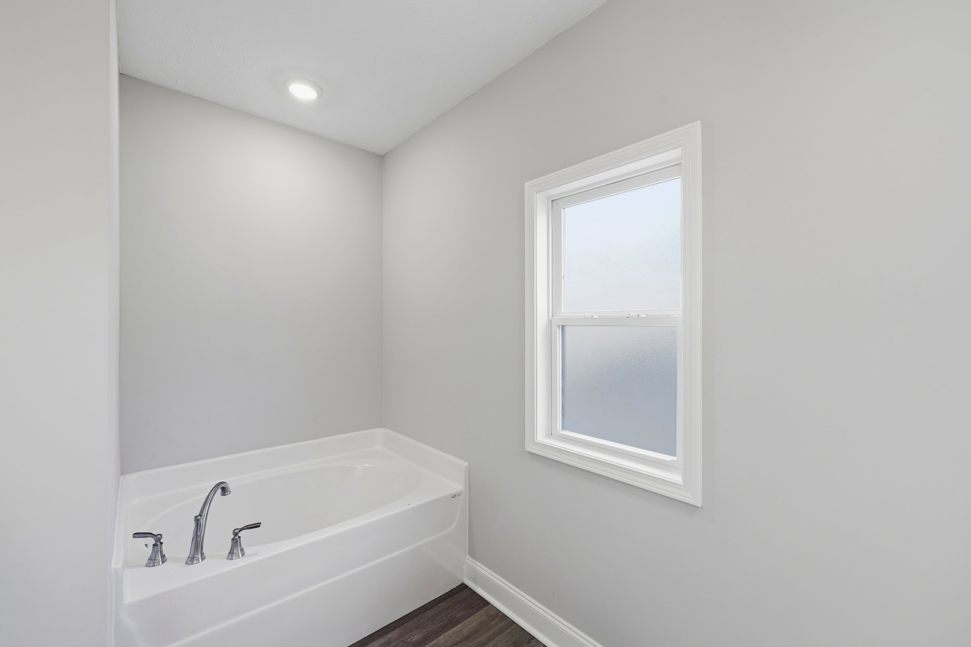 White freestanding bathtub beneath frosted glass window, chrome faucet, light gray tile flooring, plaster walls, ceiling light fixture