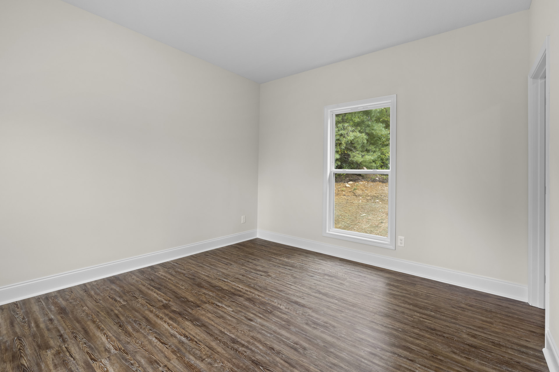 Corner of a room with light hardwood flooring, white plaster walls, and a large window overlooking a dirt field and trees.