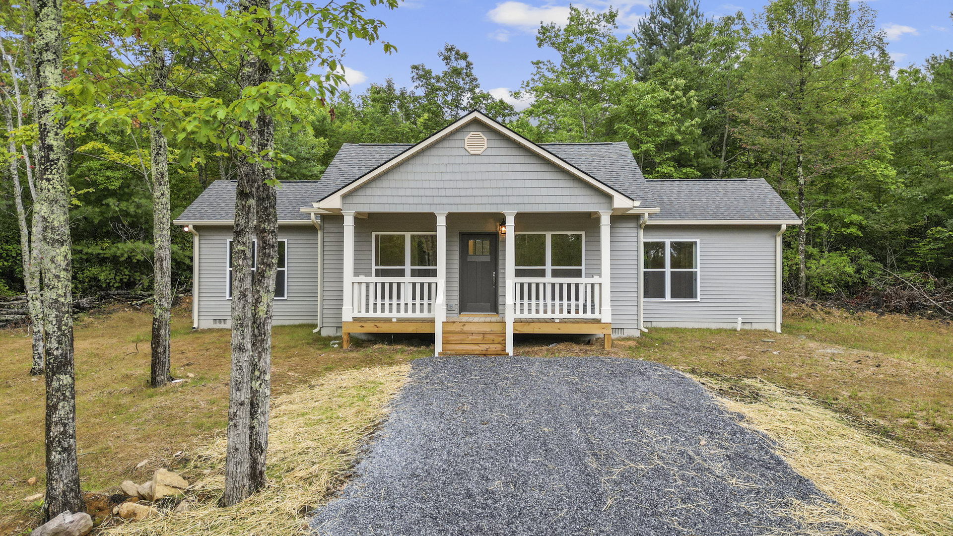 Gravel driveway bordered by grass leads to a custom home with a covered porch, white framed windows, exterior vent, and close-up view of the entry door.