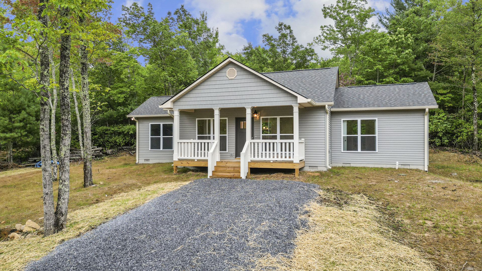 Two-story home with white-framed windows, wooden porch and stairs, white railing, gravel driveway bordered by grass, mature trees in front yard, cloudy sky overhead